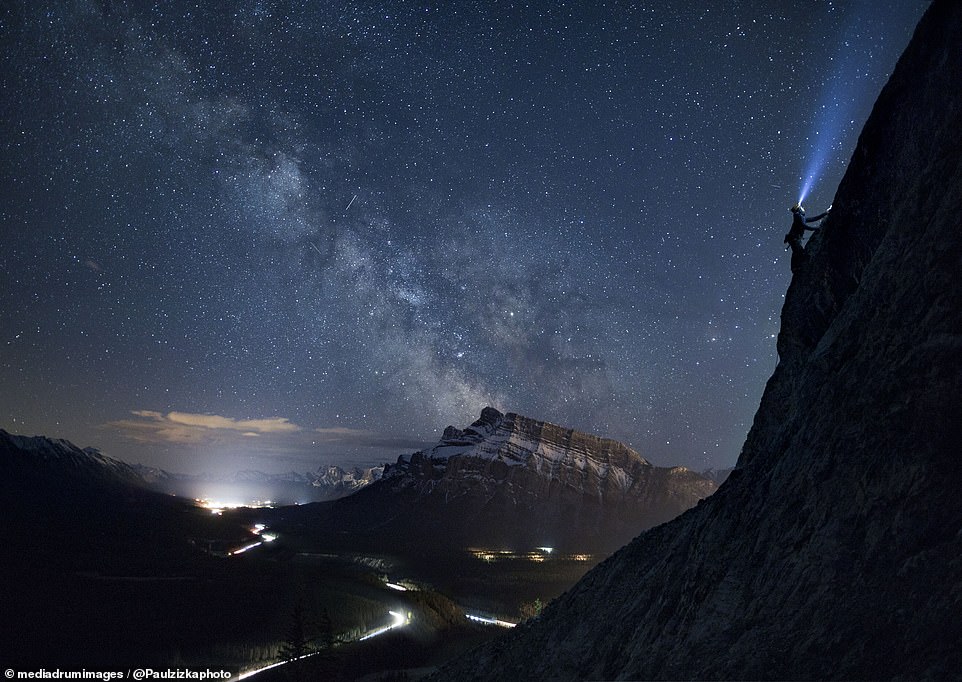 In this shot, also taken in Banff, a climber can be seen holding a flashlight as the lights of a road and houses shine below.