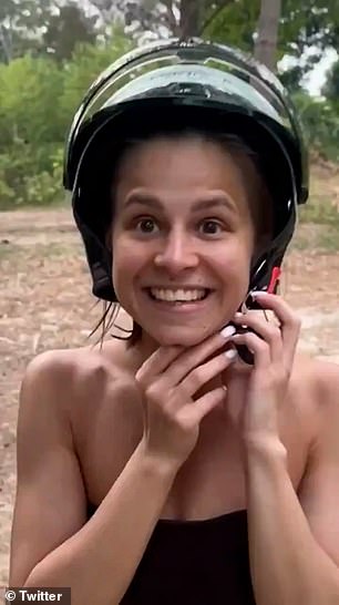 The woman can be seen smiling and whispering as she unbuckles her bicycle helmet with the elephant in the background next to a tree.