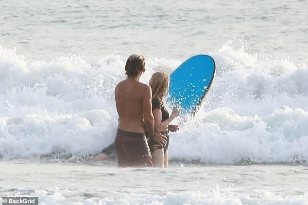 Armando places his hands on Ellie's waist as they watch a fellow surfer on the beach.