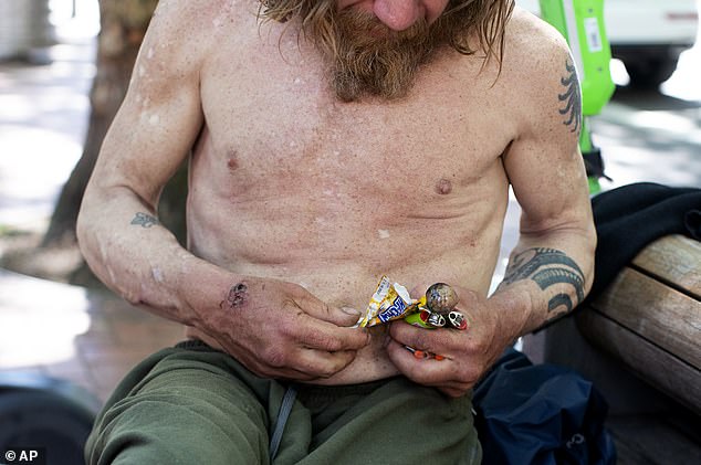 The new bill reverses a key part of the state's drug decriminalization law, which was the first of its kind in the U.S. (Pictured: A man holding a glass pipe and two lighters struggles to removing candy from a wrapper while sitting on a bench in downtown Portland)