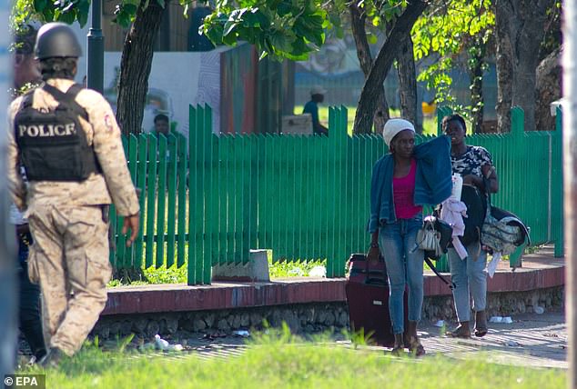 In the image: a police officer passes by while two people carry their belongings.