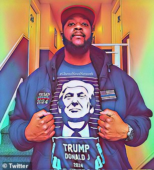 A black man is seen wearing a t-shirt with Donald Trump holding a sign and a jacket with a Trump 2024 patch.