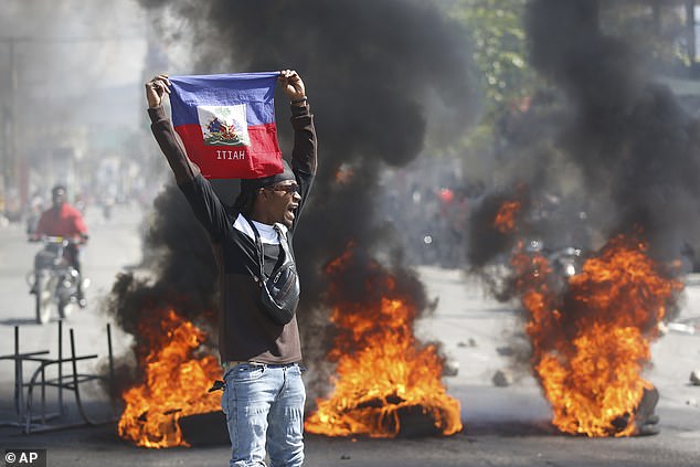 Gangs have burned down police stations, blown up the main international airport, which remains closed, and raided Haiti's two largest prisons, freeing more than 4,000 prisoners. Pictured: A protester holds a Haitian flag as tires burn in the street behind him