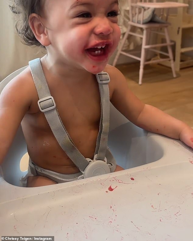 Her face, hands, and the tray of her high chair were all covered in red juice, which appeared to come from a pacifier Chrissy had handed her with cold fruit puree in the tip, possibly to soothe tooth pain.