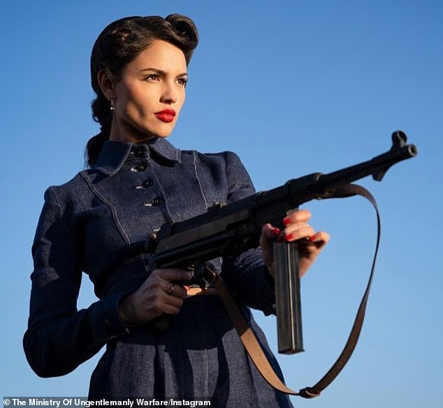 The 34-year-old actress is seen wearing a dark buttoned-up suit with her hair slicked back while holding a large gun