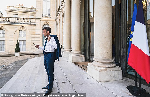 Macron is seen holding his blue jacket over his shoulder while checking his phone.