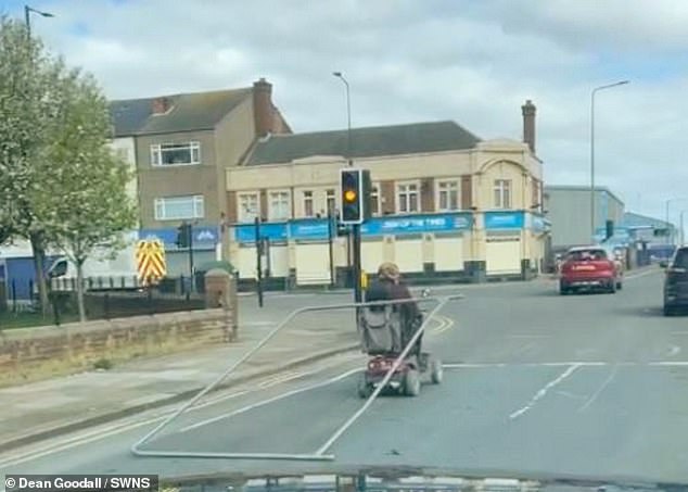 The video shows the man driving down the middle of a road on his scooter with a large metal fence tied to the headrest with a rope.