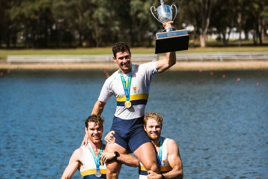 Two rowers carry another rower on their shoulders.  The rower on his shoulders raises a trophy. 