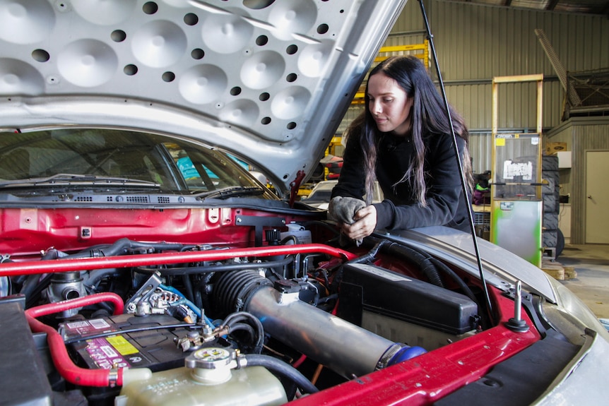 A young woman looks at the engine of a car with the hood raised.