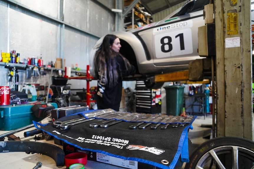 In the foreground, a woman fixes a car with mechanic tools.