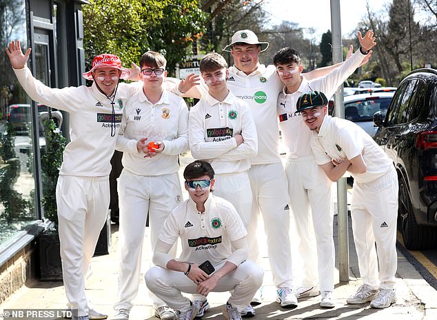 A group of boys smile while dressed in full cricket gear.  Some have sunscreen on their faces.