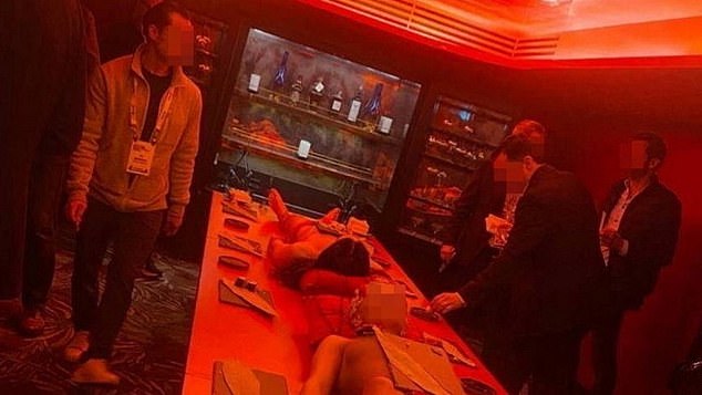 The image shows a group of men in the red lacquered room of the five-star Mandrake hotel in central London, with a partially naked man and woman lying motionless on the table in front of them.