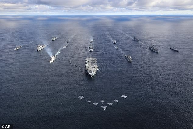 The Royal Navy's aircraft carrier HMS Prince of Wales leads a formation of fifteen ships as aircraft pass at sea for exercise Nordic Response 24