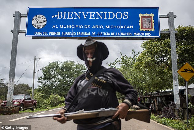 An armed member of Pueblos Unidos poses in front of the Welcome sign to the municipality of Ario de Rosales