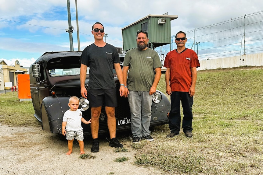 A little boy, his father, his grandfather, and his great-grandfather are standing in front of a dark-colored hot rod.