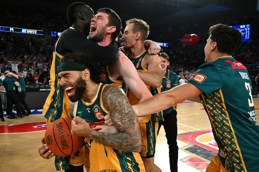 A group of Tasmanian NBL players cheer, cry and hug each other on the pitch after winning the title. 
