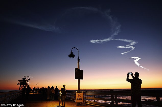 People walk on a pier beneath the wake of a SpaceX Falcon 9 rocket in San Clemente