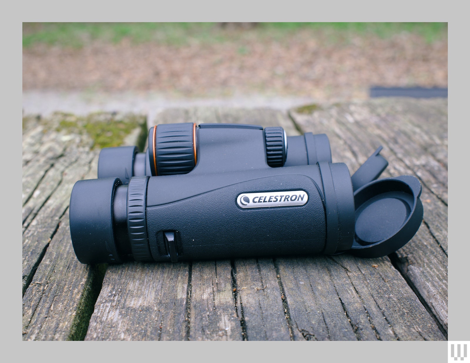 Side view of a pair of black binoculars with the lens caps partially loose, sitting on a wooden surface with dry leaves in the background
