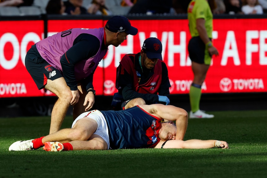 A Melbourne AFL player lies injured on the ground as two medical staff watch closely.