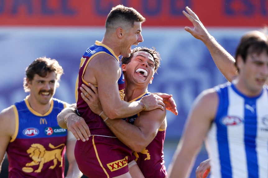 Two Brisbane Lions players shout and smile in celebration after a goal, as a Kangaroos defender walks away.