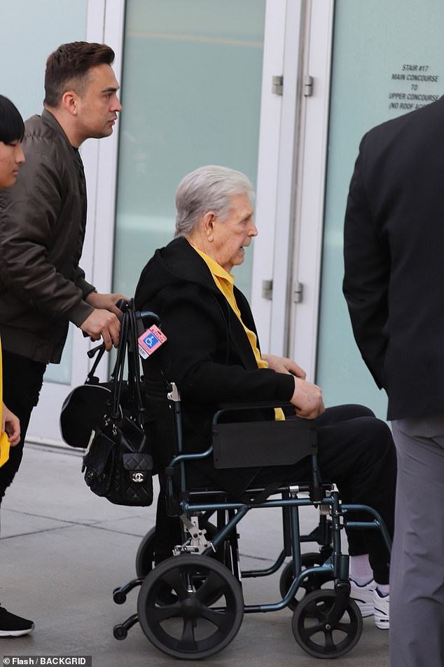 He supported his team with a yellow polo shirt and also a black fleece jacket, black sweatpants and black and white Nike sneakers. A man pushed Wilson's wheelchair, while a woman rested a hand on his shoulder as they headed toward a door to enter the arena.