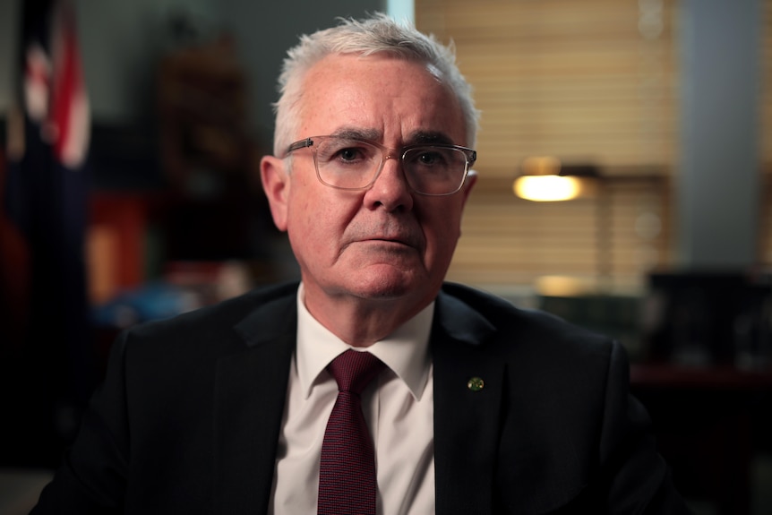 A middle-aged man wearing glasses, a suit and tie, is sitting in his office looking at the camera.  Behind him is an Australian flag.