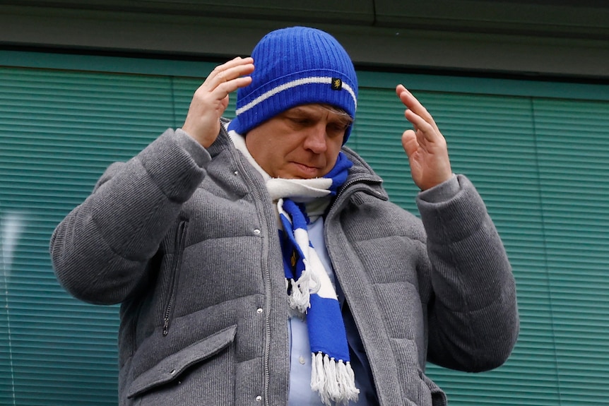 A man wearing a blue hat and a blue and white scarf gestures with his hands