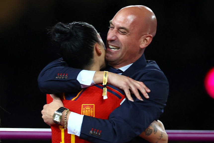 A smiling man dressed in a suit hugs a woman dressed in a soccer uniform