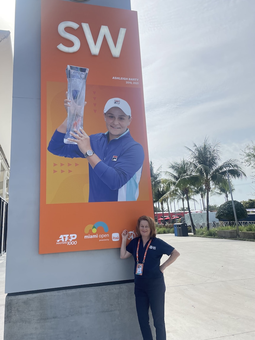 Deirde stands next to a large sign with Ash Barty holding a trophy, pointing at the sign in a blue polo shirt.