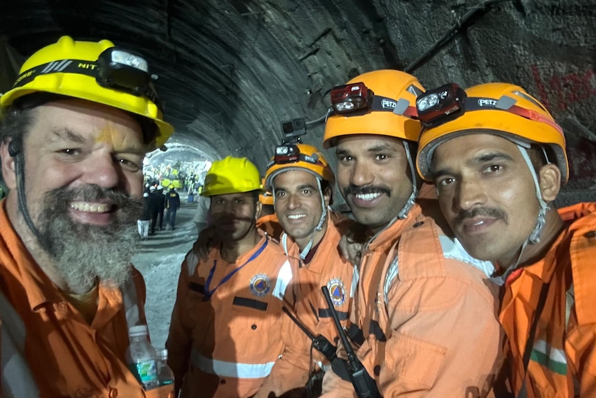 A group of miners in orange high-visibility gear and helmets smile inside a dark tunnel