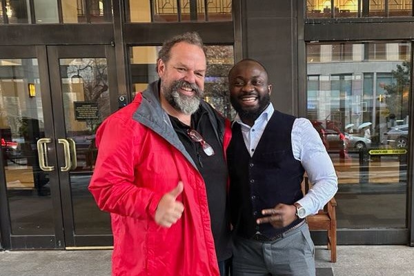 Two men stand next to each other, one in a red jacket and beard, the other in a business suit, smiling 
