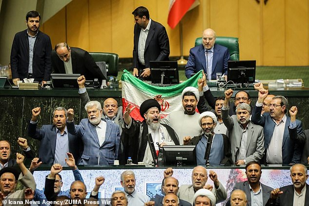 Iranian parliamentarians gathered in parliament punching the air and chanting victoriously as the Iranian flag rises above them.