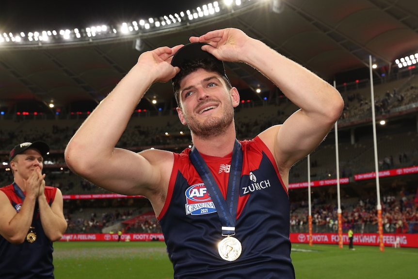 A smiling Melbourne AFL player wears a cap and a medal around his neck after his team won a grand final. 