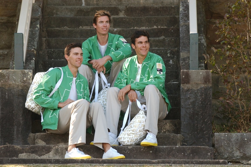 Three male athletes sitting on steps wearing gray pants and green jackets with white outlines. 