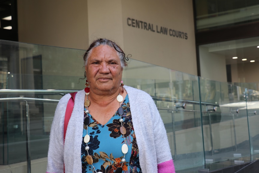 An older Indigenous woman in a blue floral top and gray cardigan posing for a photograph outside the court in Perth.