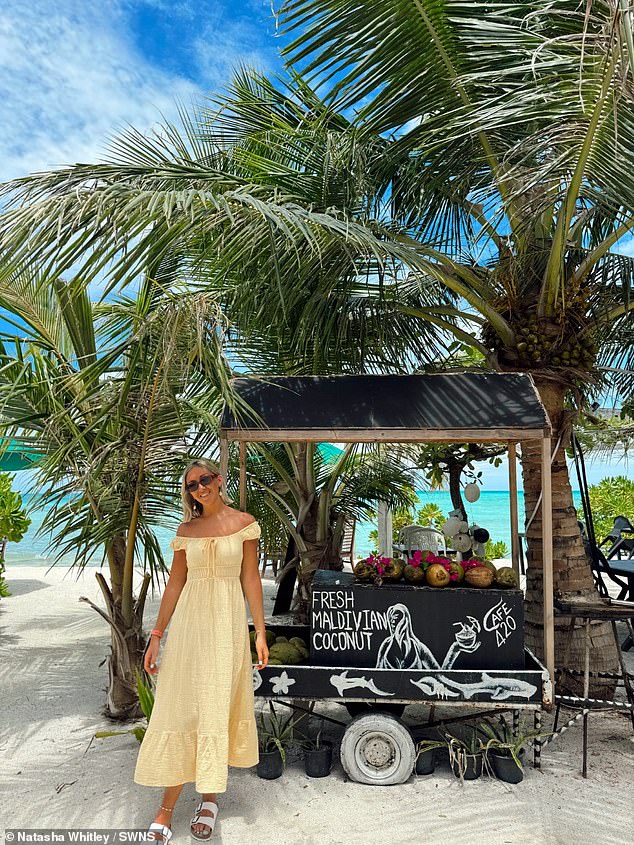 Natasha, wearing a summery yellow dress, smiles for a photo next to a fresh coconut stall in Dhiffushi
