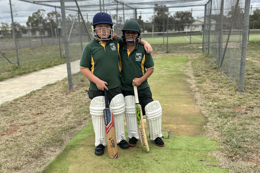 Two boys in school green brief, cricket pads, helmets and with a bat hug each other in cricket nets.