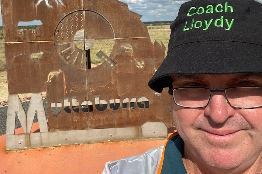A guy with glasses and a "Coach Lloydy" Bucket hat stands in front of a rusty Qld metal sculpture with the word Muttaburra 