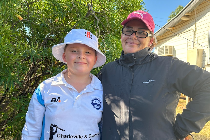 A boy in cricket gear and a mother in a jacket, red cap and reading glasses.