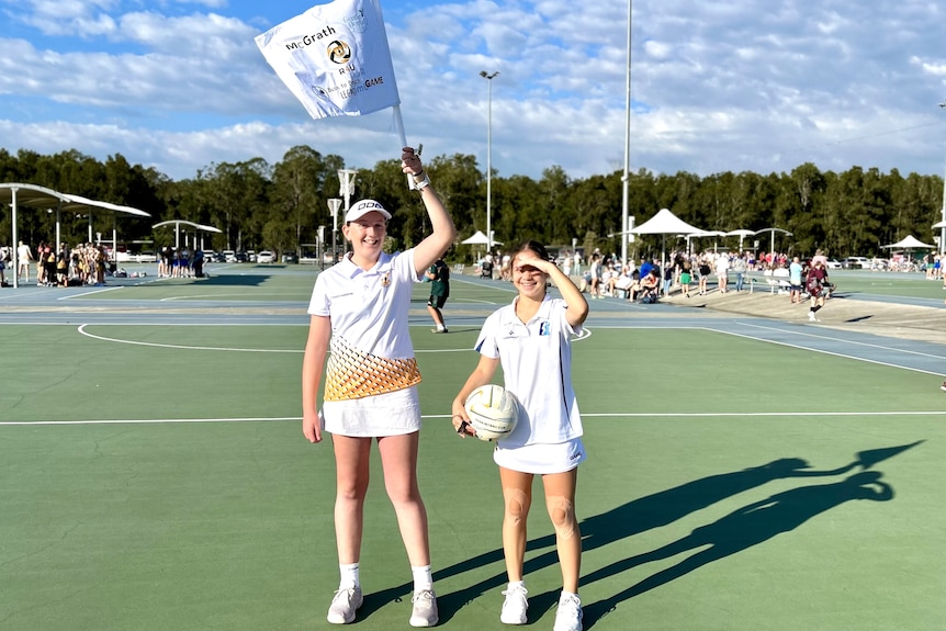 Two girls dressed in white, standing on a netball court.  One holds a white flag over her head.