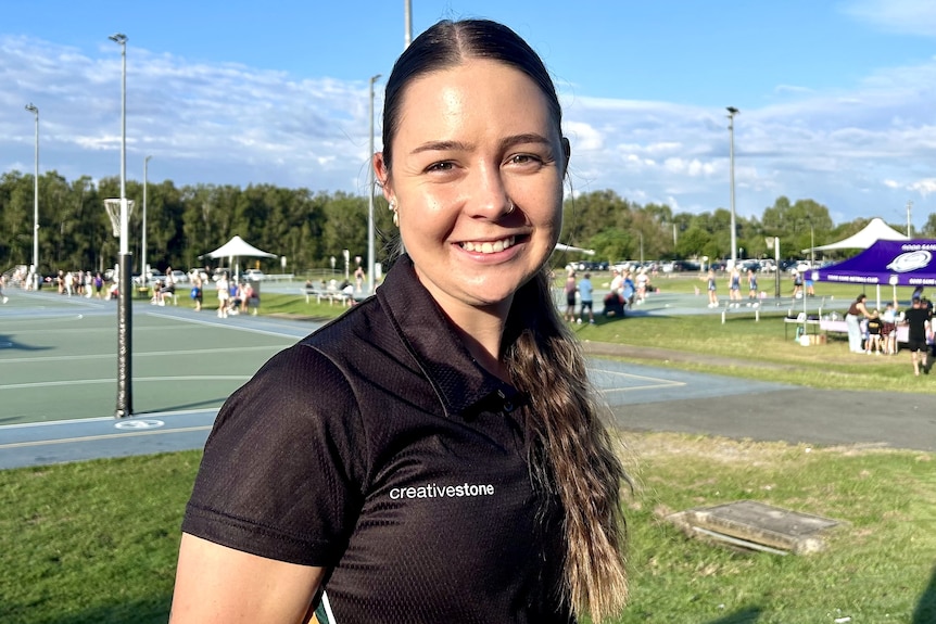 A young woman with long dark hair tied back and wearing a dark shirt.  She is standing near a netball court, smiling.