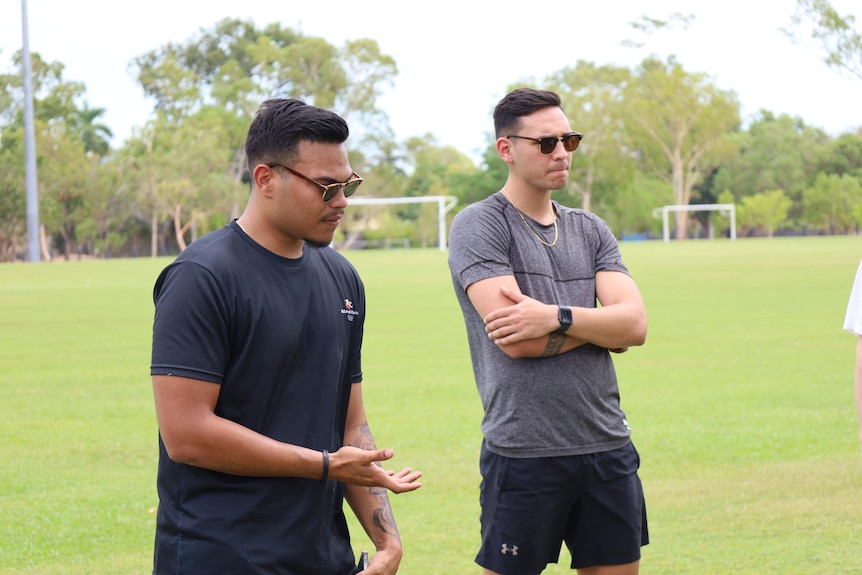 Two men appear in the photo, both wearing sunglasses and dark shirts.  They are standing on a field with soccer goals.