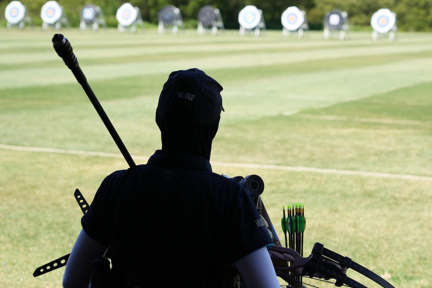 An archer is shown in the shadows at one end of a shooting range, with a row of archery targets in the distance.