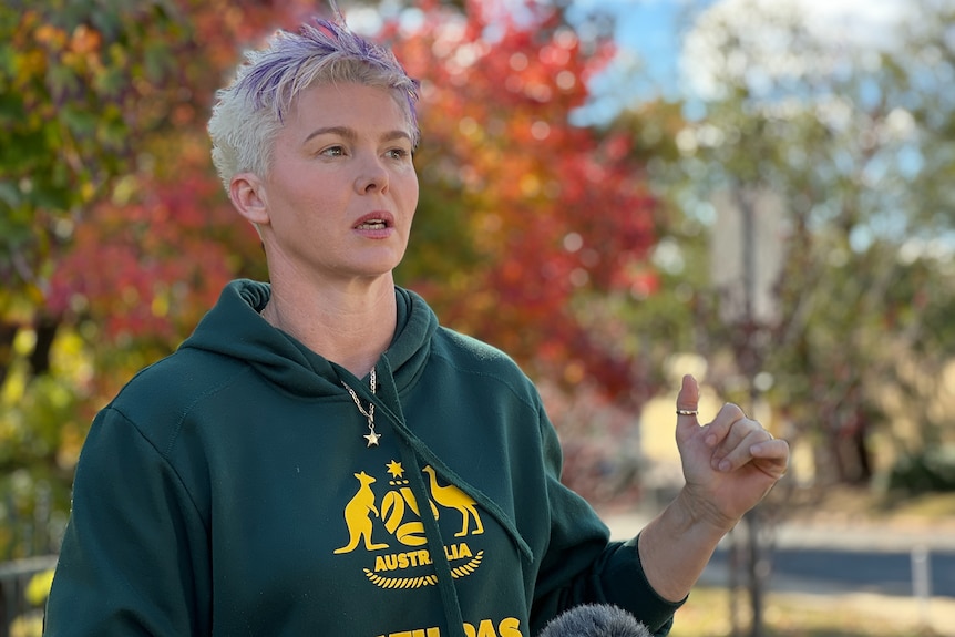 A short-haired woman in a Matildas hoodie speaks in front of a microphone