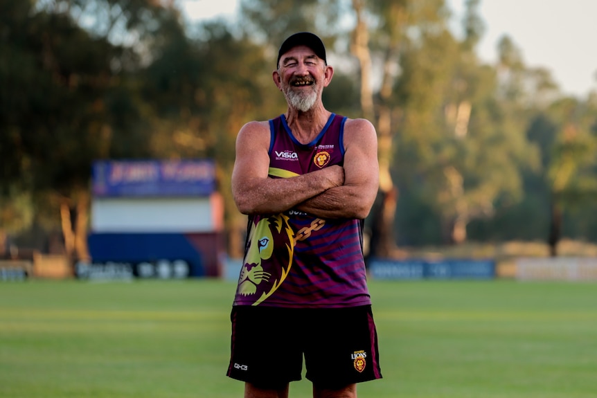 An older man in a cap, t-shirt and shorts laughs while standing at the national football oval