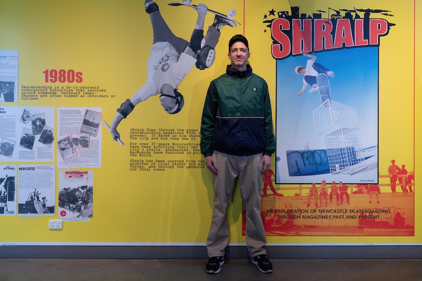 A man wearing a green jacket stands in front of a yellow wall covered in old magazine covers of people skating.