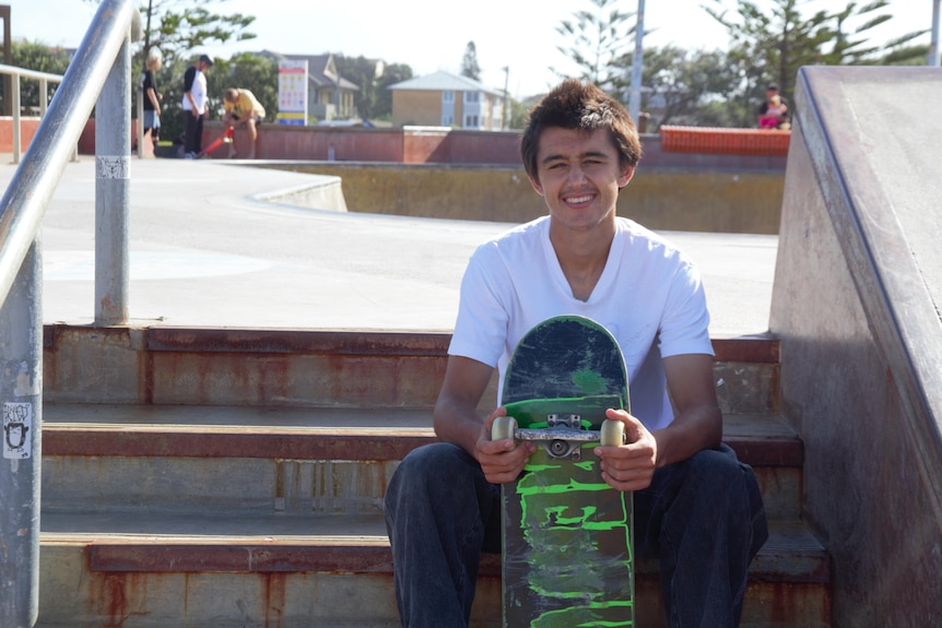 A young man in a white shirt is sitting on some concrete steps in front of a skate bowl smiling.  He is holding his skateboard in his hands.