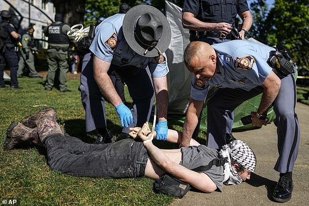 A protester wearing a keffiyeh is seen pinned to the ground while officers put zip ties on his wrist.