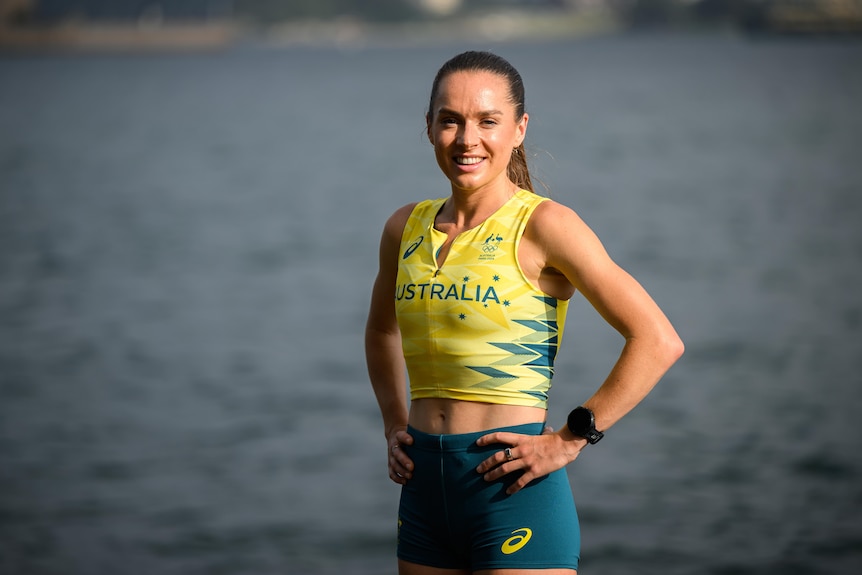 A runner, Izzi Batt-Doyle, wears the uniform of the Australian Olympic team, poses with her hands on her hips and smiles.