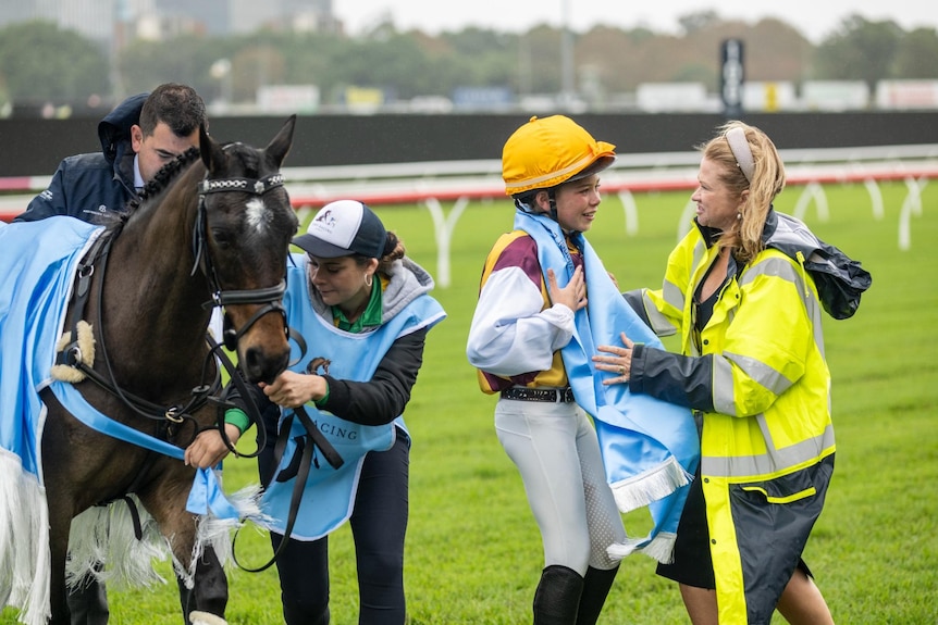 A woman puts a scarf on a girl dressed as a jockey next to a horse